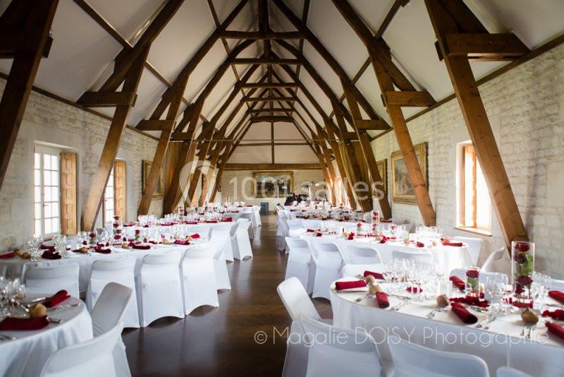 Salle de réception lumineuse avec des tables rondes dressées, nappes blanches et décorations rouges, poutres apparentes.