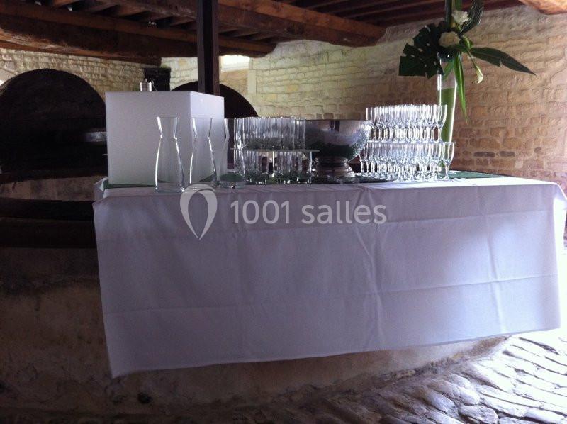 Table dressée avec une nappe blanche, contenant des verres, carafes et une coupe, dans un cadre en pierre rustique.