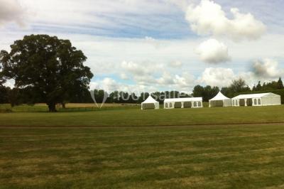 Grand espace vert avec des tentes blanches installées sur une pelouse, entouré d'arbres et d'un ciel partiellement nuageux.