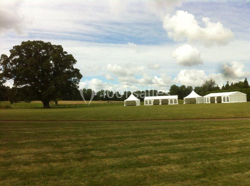 Grand espace vert avec des tentes blanches installées sur une pelouse, entouré d'arbres et d'un ciel partiellement nuageux.
