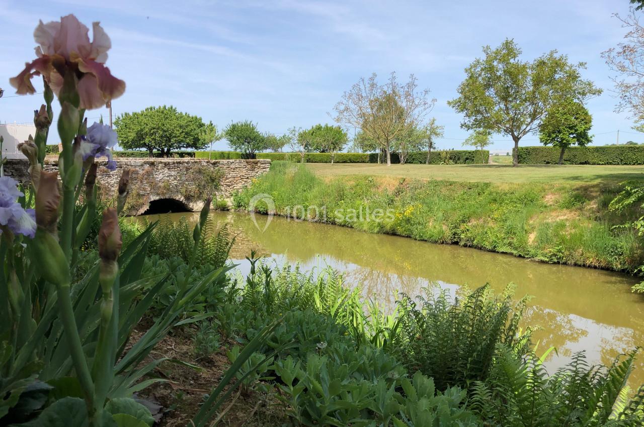 Vue d'un petit cours d'eau bordé de végétation, avec un pont en pierre et des arbres sous un ciel dégagé.