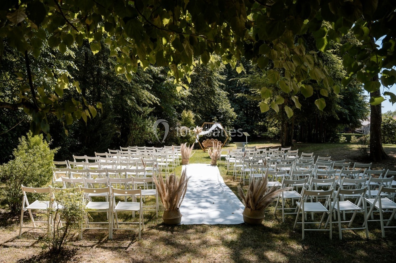 Allée centrale décorée pour une cérémonie de mariage en extérieur, entourée de chaises blanches et de verdure.