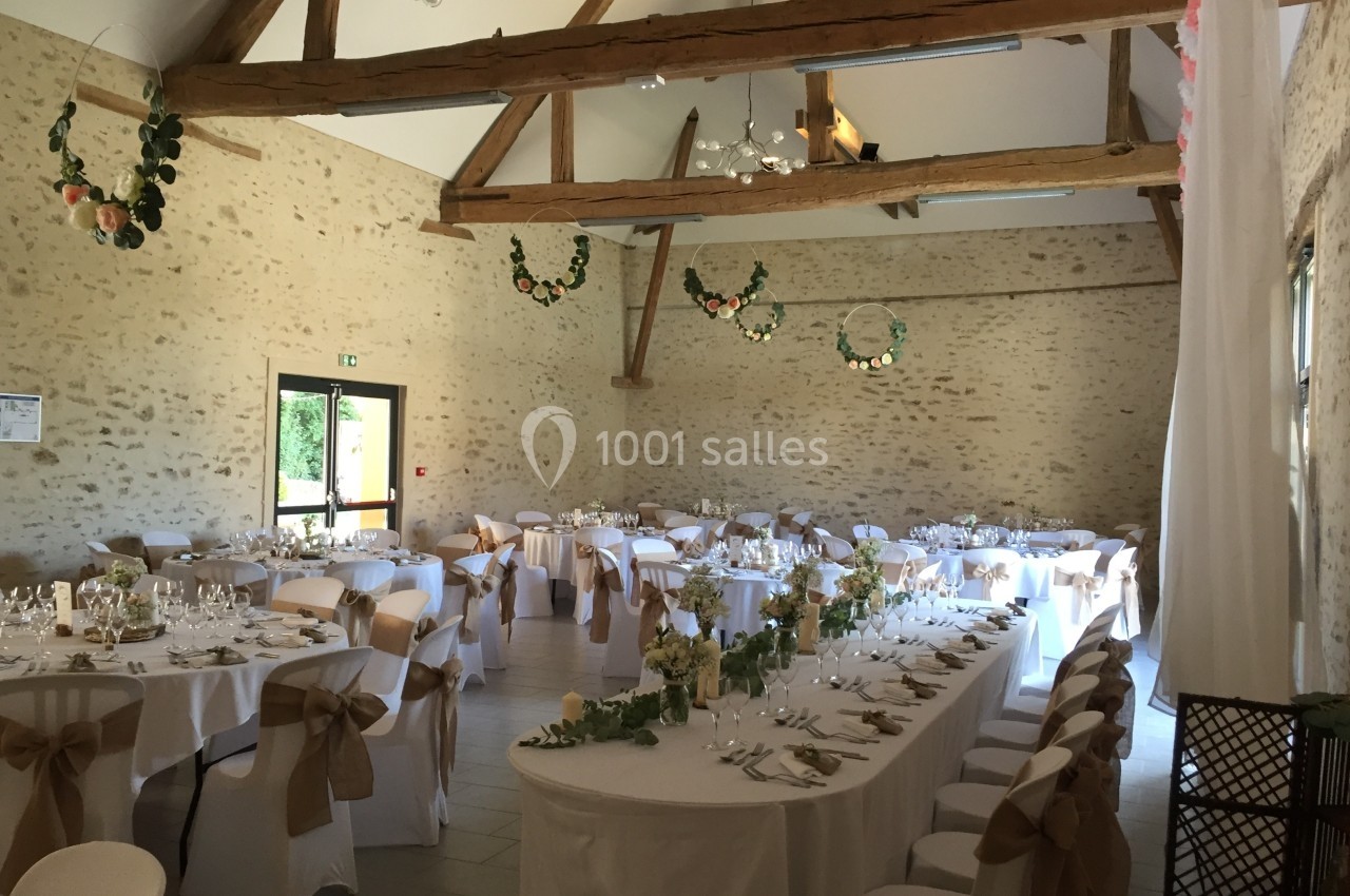 Salle de réception lumineuse avec tables décorées, nappes blanches et poutres apparentes en bois.
