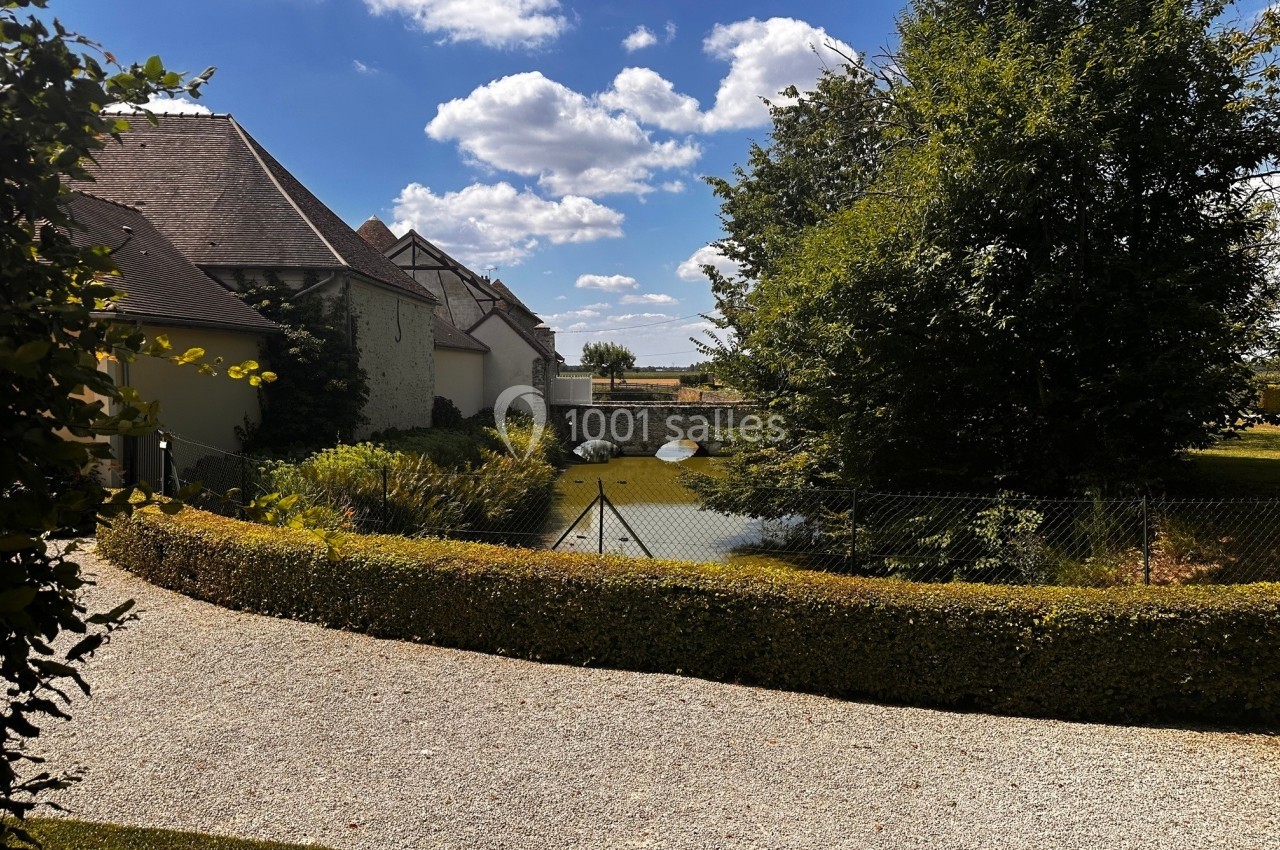 Chemin gravillonné bordé de haies menant à un étang entouré de végétation et de bâtiments sous un ciel bleu.