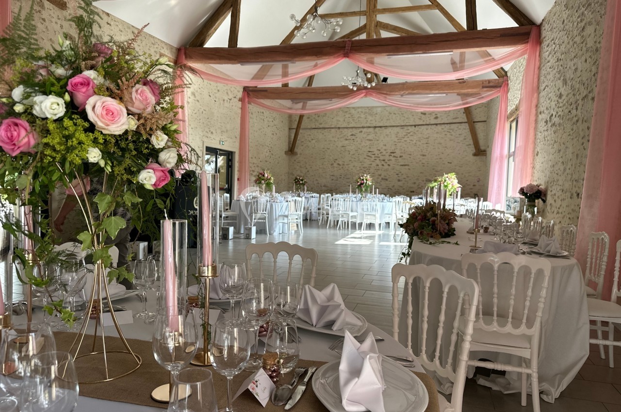 Salle de réception décorée avec des fleurs roses, nappes blanches et chaises, sous une charpente en bois apparente.