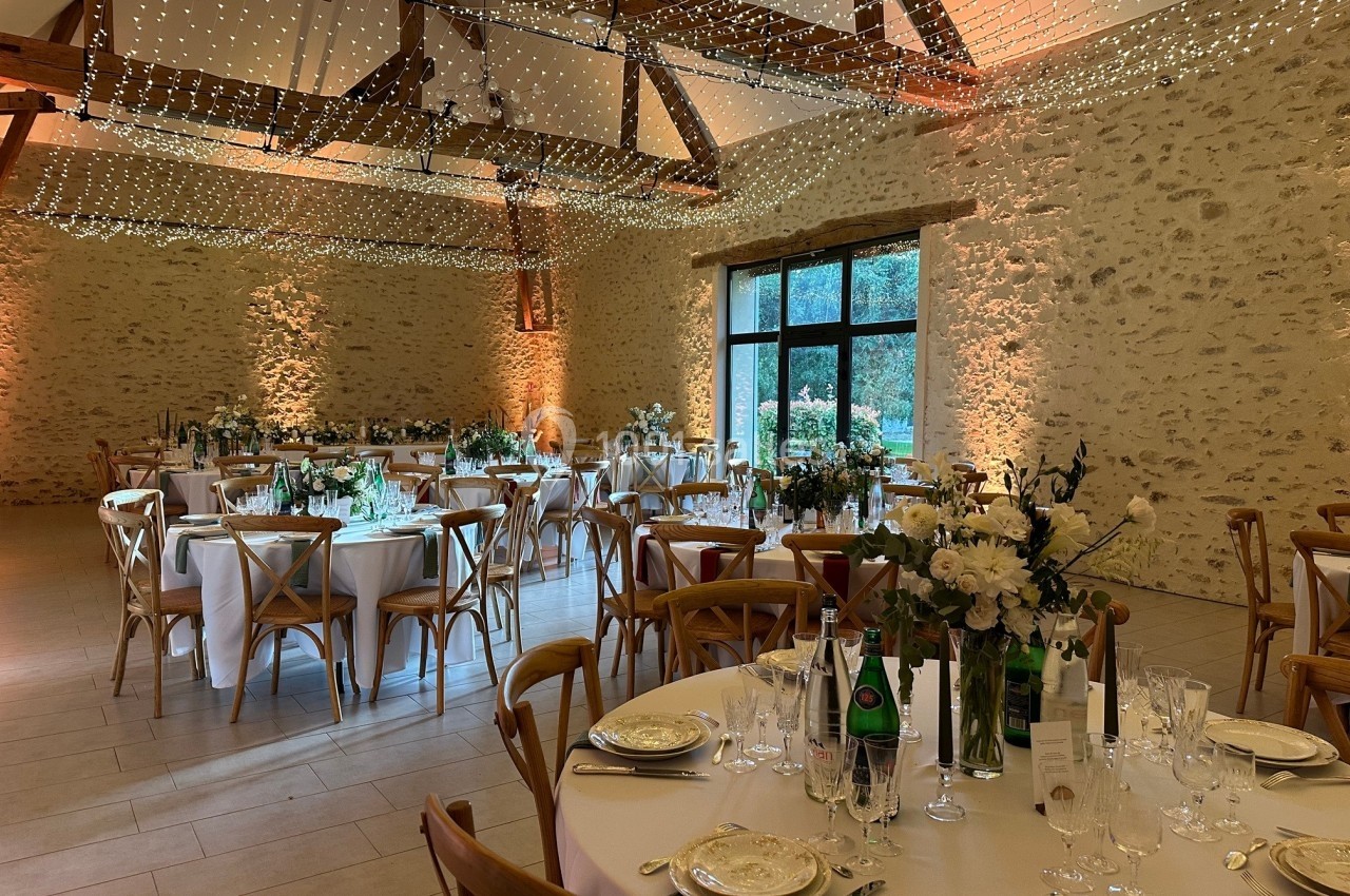 Salle de réception décorée avec des tables rondes dressées, des chaises en bois et un plafond illuminé de guirlandes…