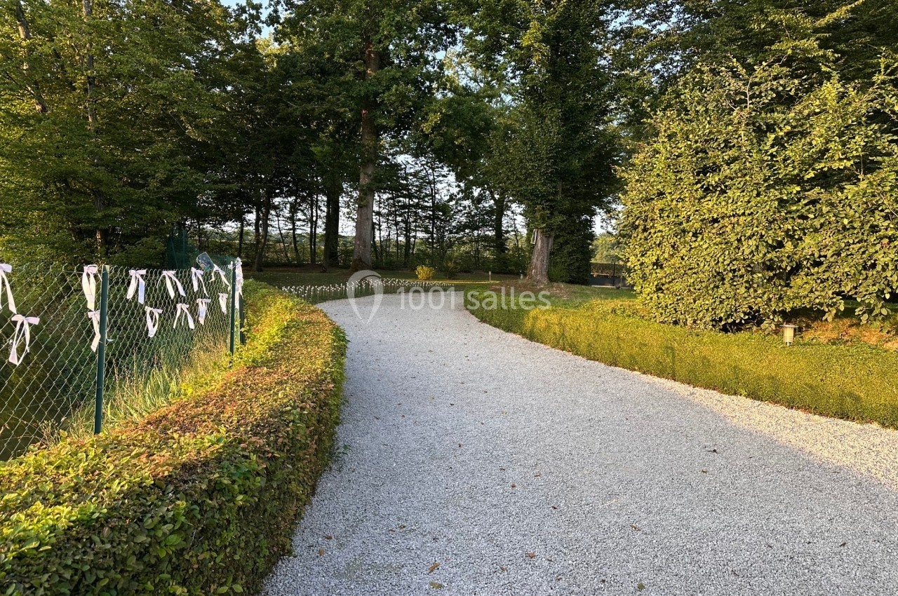 Allée de gravier bordée de haies taillées et d'arbres, avec des rubans blancs accrochés à un grillage sur la gauche.