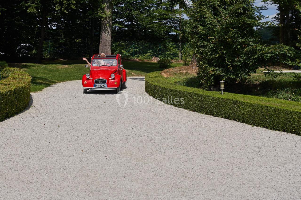 Voiture rouge ancienne roulant sur une allée gravillonnée bordée de haies dans un cadre verdoyant.