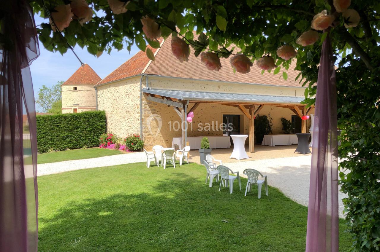 Vue d'une cour verdoyante avec une grange en pierre, des chaises blanches et des tables hautes décorées.