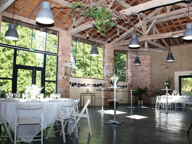 Salle lumineuse avec grandes fenêtres, poutres apparentes, tables dressées et décoration végétale suspendue.