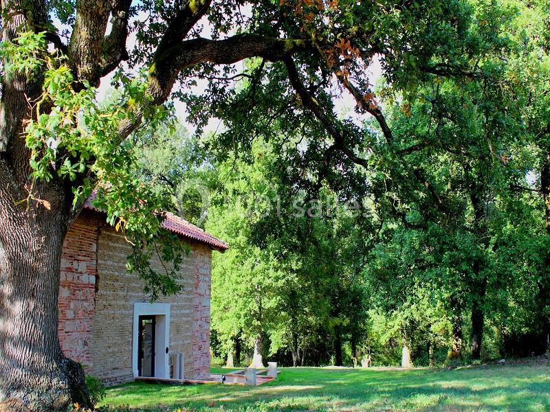Bâtisse en pierre avec toit rouge entourée d'arbres dans un espace verdoyant et ensoleillé.