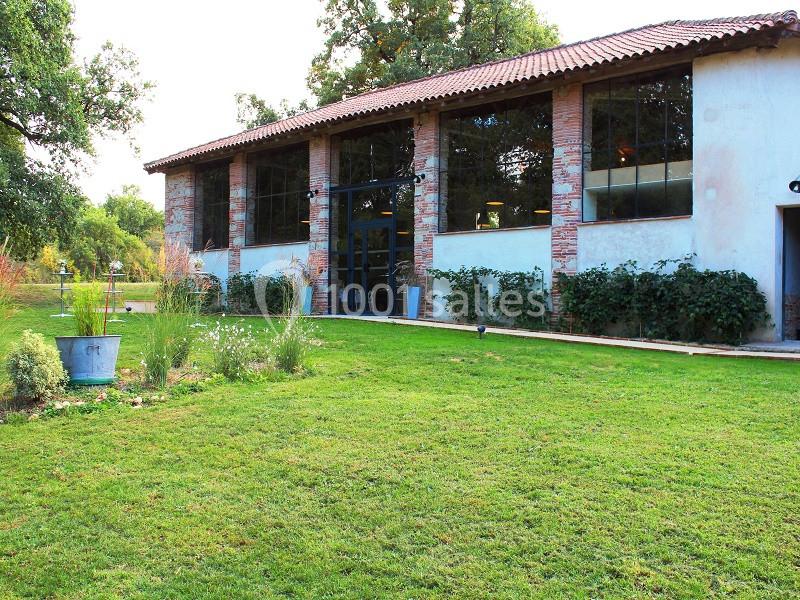 Bâtiment en pierre avec de grandes fenêtres, entouré d'un jardin verdoyant et d'arbustes.