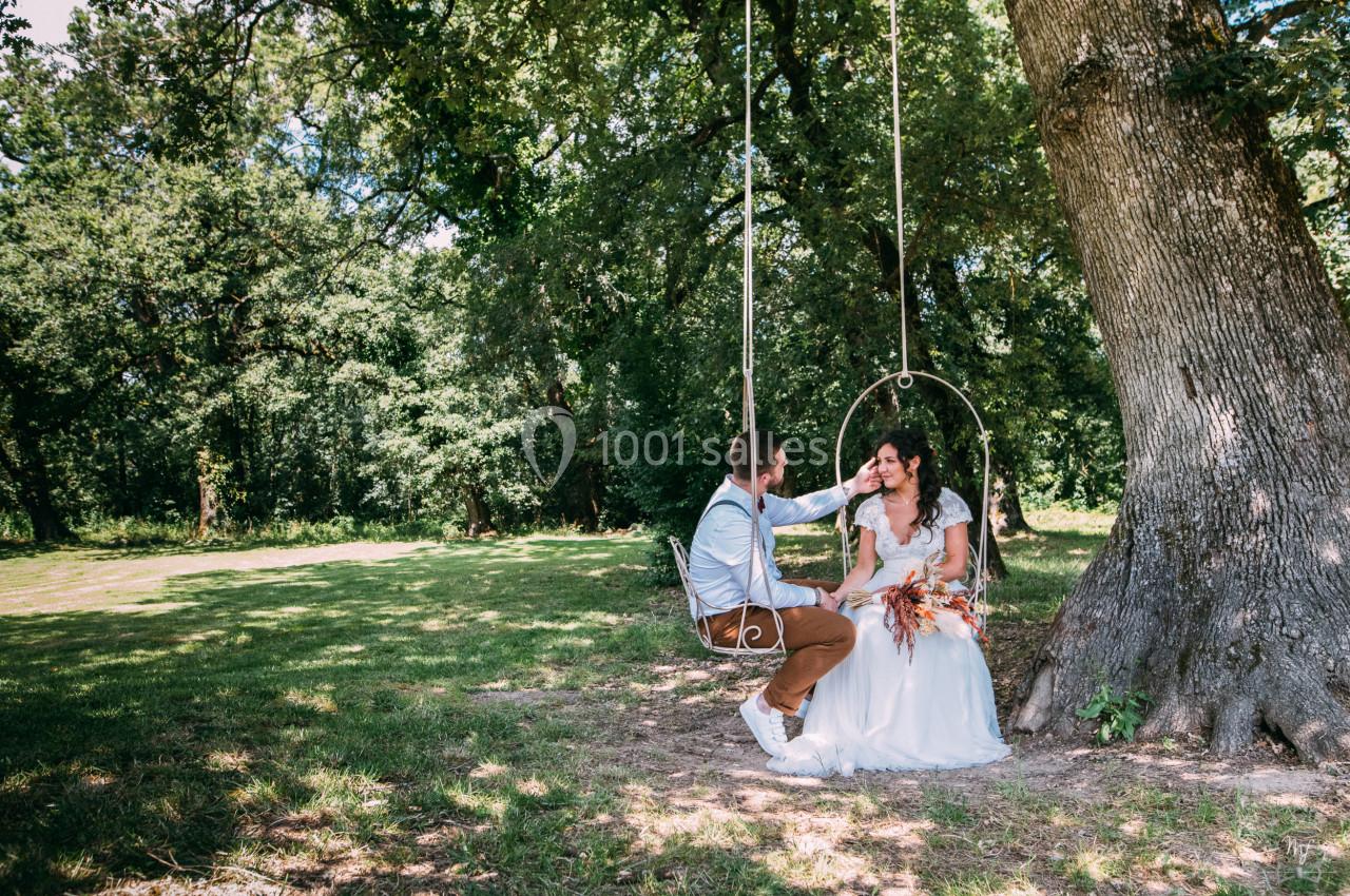 Un couple assis sur une balançoire suspendue à un arbre dans un parc verdoyant, échangeant un moment complice.