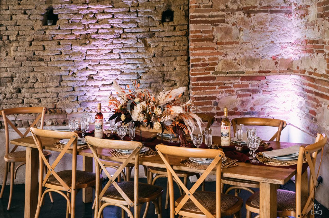 Table en bois dressée avec des assiettes, verres et un centre de table floral, devant un mur en briques éclairé.