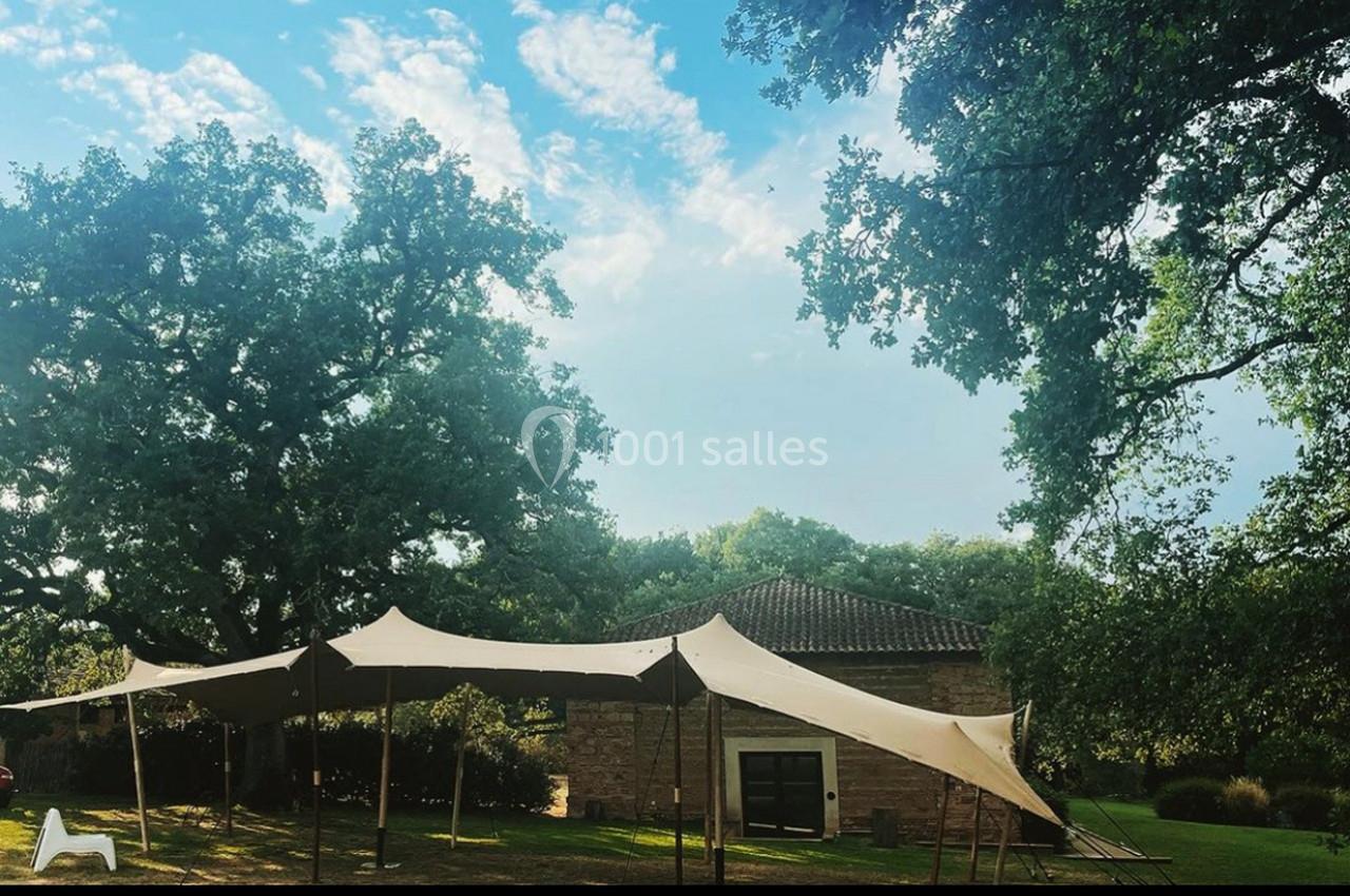 Tente beige installée dans un jardin arboré, près d'un bâtiment en pierre sous un ciel partiellement nuageux.