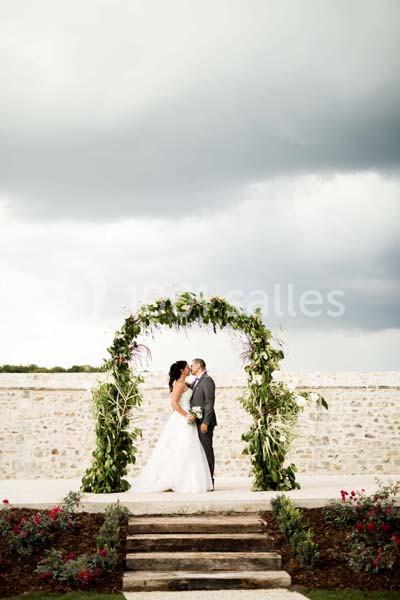 Un couple de mariés s'embrasse sous une arche végétale, avec un mur en pierre et un ciel nuageux en arrière-plan.