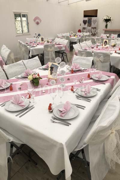Salle décorée pour un événement avec des tables dressées, nappes blanches, chemins de table roses et pliages de serviettes.