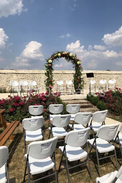 Chaises blanches disposées en rangées devant une arche fleurie dans un jardin sous un ciel ensoleillé.