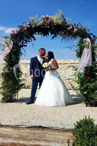 Un couple de mariés s'embrasse sous une arche fleurie en extérieur par une journée ensoleillée.