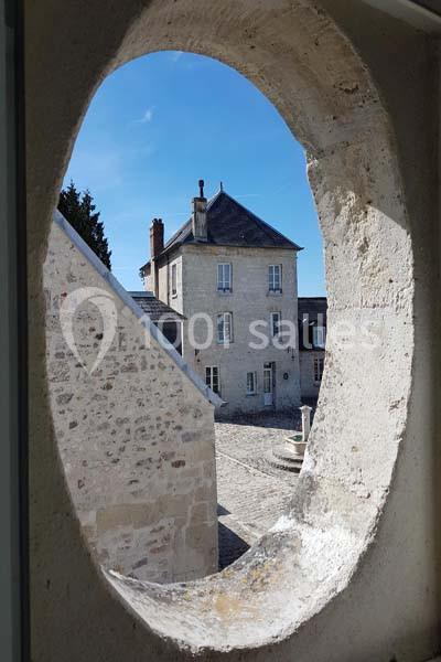 Vue d'une cour pavée et d'une maison en pierre, encadrée par une ouverture ovale dans un mur.