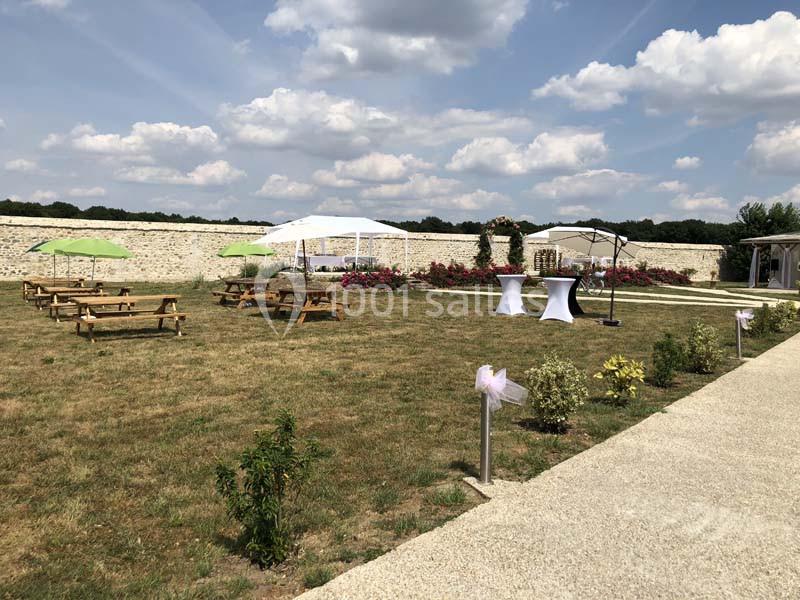 Espace extérieur avec pelouse, tables en bois, tonnelles blanches et décorations florales sous un ciel partiellement nuageux.