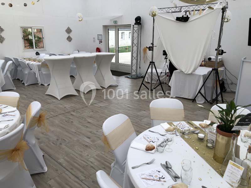 Salle de réception décorée avec des tables rondes dressées, nappes blanches et éléments dorés, espace photo en arrière-plan.