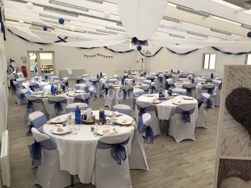 Salle de réception décorée pour un événement, avec des tables rondes dressées et des chaises ornées de nœuds bleus.