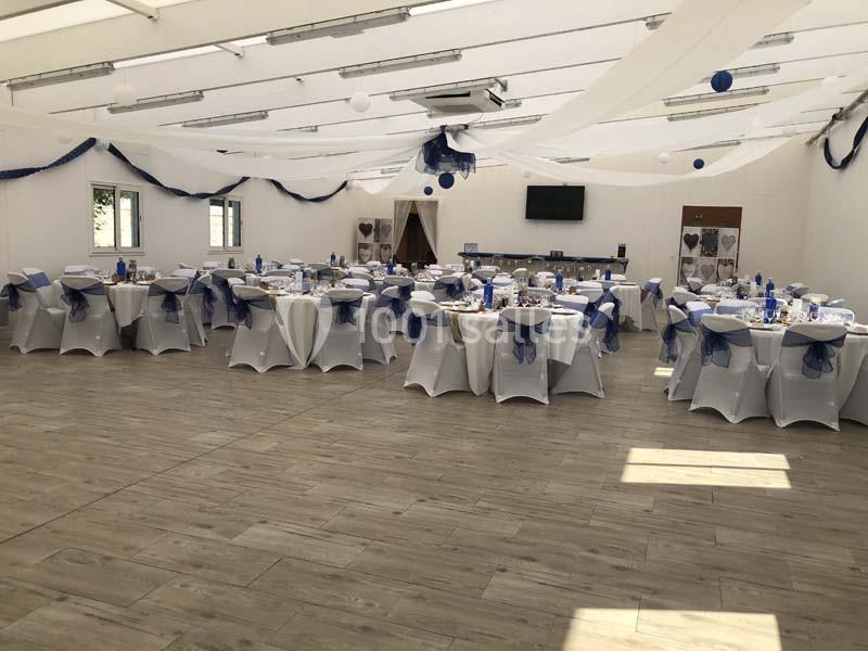 Salle de réception décorée avec des tables rondes, nappes blanches et nœuds bleus, sous des guirlandes suspendues.