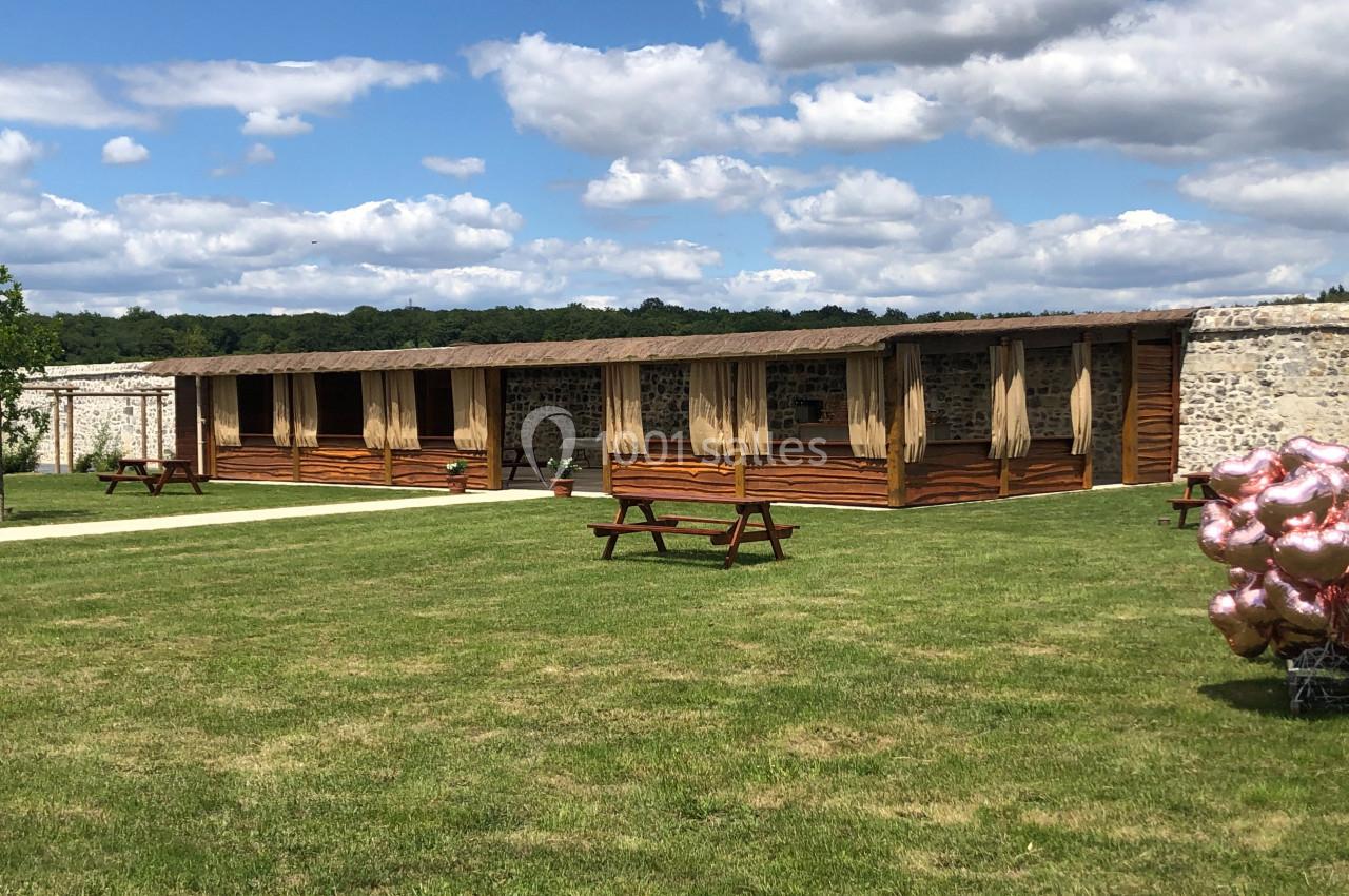 Un bâtiment en bois avec des rideaux ouverts, entouré d'une pelouse et de quelques bancs sous un ciel partiellement nuageux.
