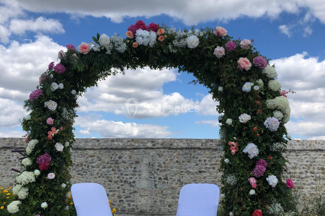 Arche fleurie ornée de roses et pivoines avec deux chaises blanches, installée en extérieur sous un ciel nuageux.