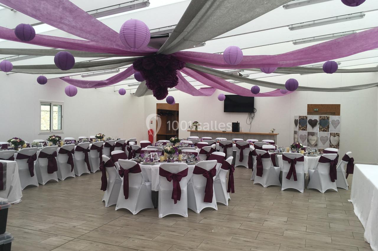 Salle de réception décorée avec des nappes et des rubans violets, des tables dressées et des lanternes suspendues.