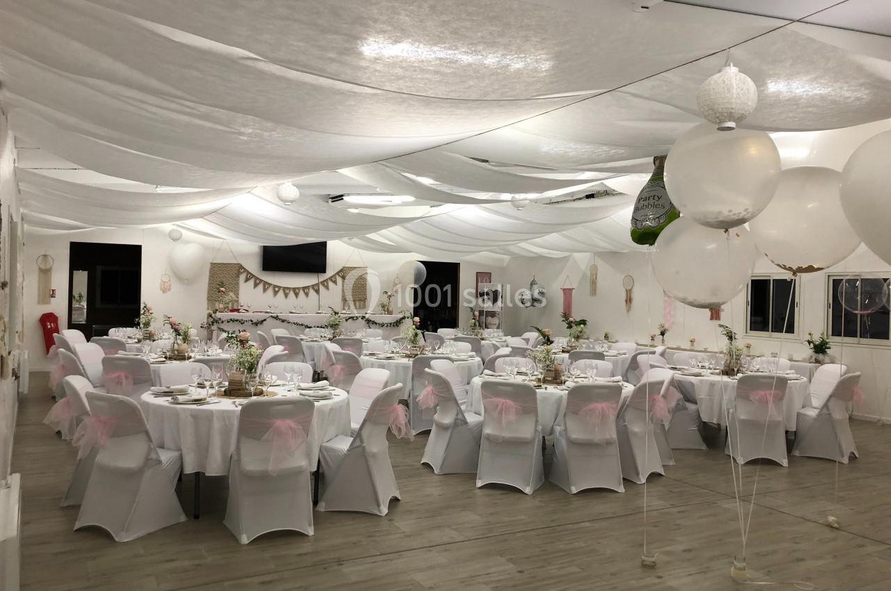 Salle de réception décorée avec des tables rondes, nappes blanches, chaises couvertes et ballons suspendus.