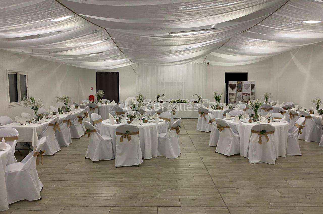 Salle de réception décorée avec des tables rondes, nappes blanches, chaises ornées de rubans dorés et arrangements floraux.