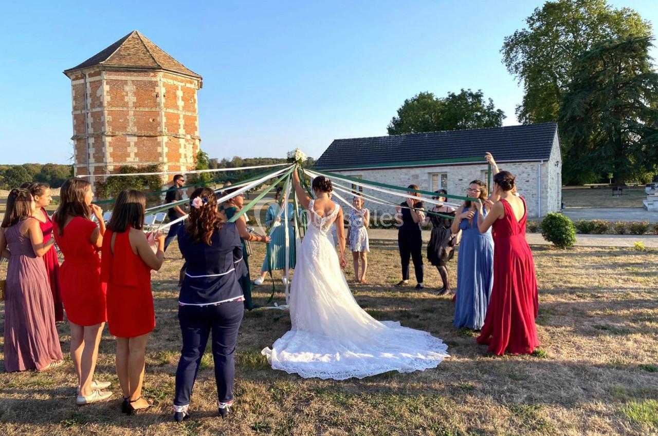 Un groupe de personnes en cercle tient des rubans autour d'une mariée, dans un jardin près de bâtiments anciens.