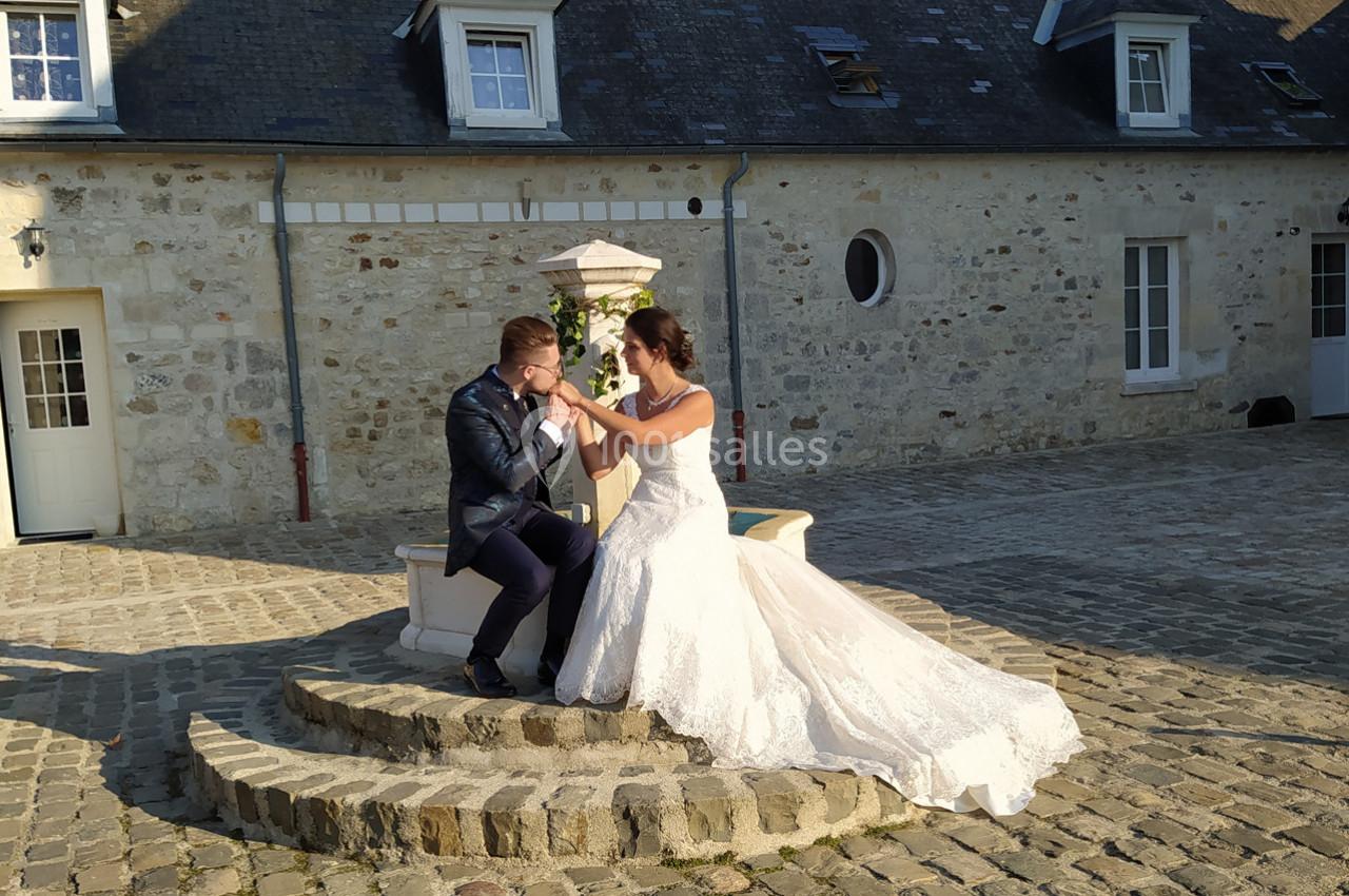 Un couple en tenue de mariage assis sur un muret en pierre dans une cour pavée devant un bâtiment en pierre.