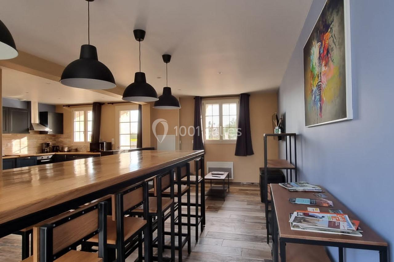 Salle à manger lumineuse avec une grande table en bois, des chaises noires, des suspensions et un mur décoré d'un tableau…