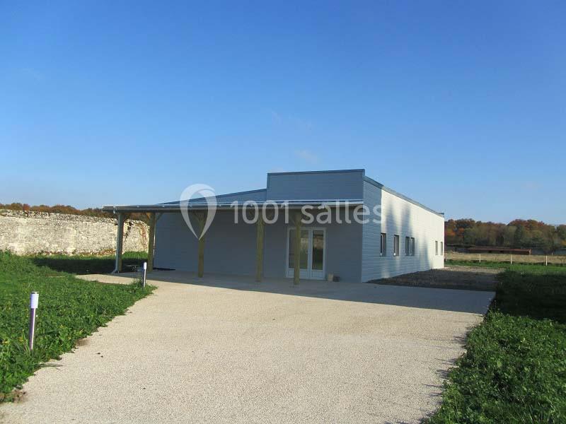 Bâtiment moderne de plain-pied avec façade grise, abri en bois et allée en gravier, entouré de verdure.