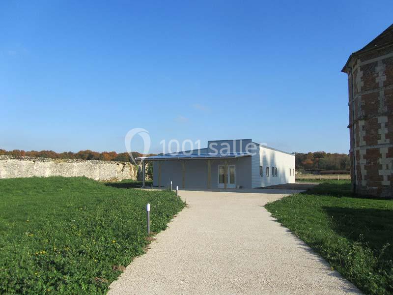 Bâtiment moderne avec façade claire, situé au bout d'une allée gravillonnée, entouré de verdure et de murs anciens.