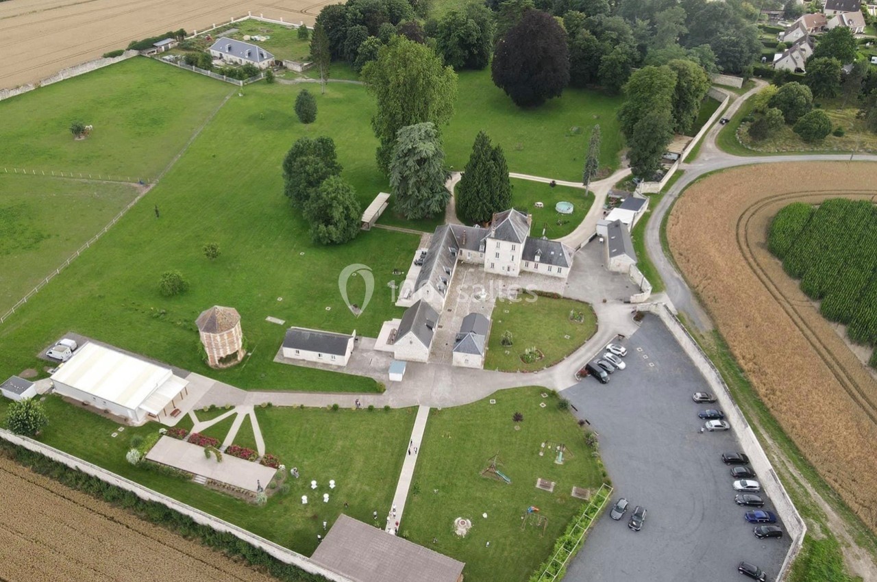 Vue aérienne d'un domaine rural avec bâtiments, espaces verts, parking et champs environnants.