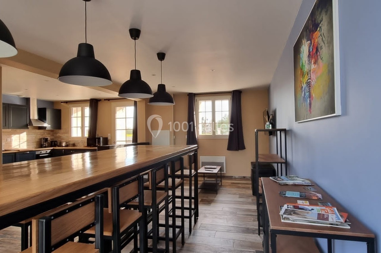 Salle à manger lumineuse avec une grande table en bois, des chaises hautes, des suspensions noires et un coin lecture.