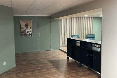 Couloir avec parquet, murs verts, porte au fond, porte-manteaux vides, bancs et panneau indiquant des toilettes.