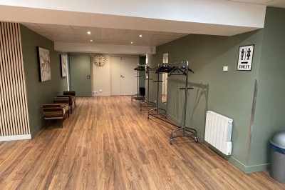 Couloir avec parquet, murs verts, porte au fond, porte-manteaux vides, bancs et panneau indiquant des toilettes.