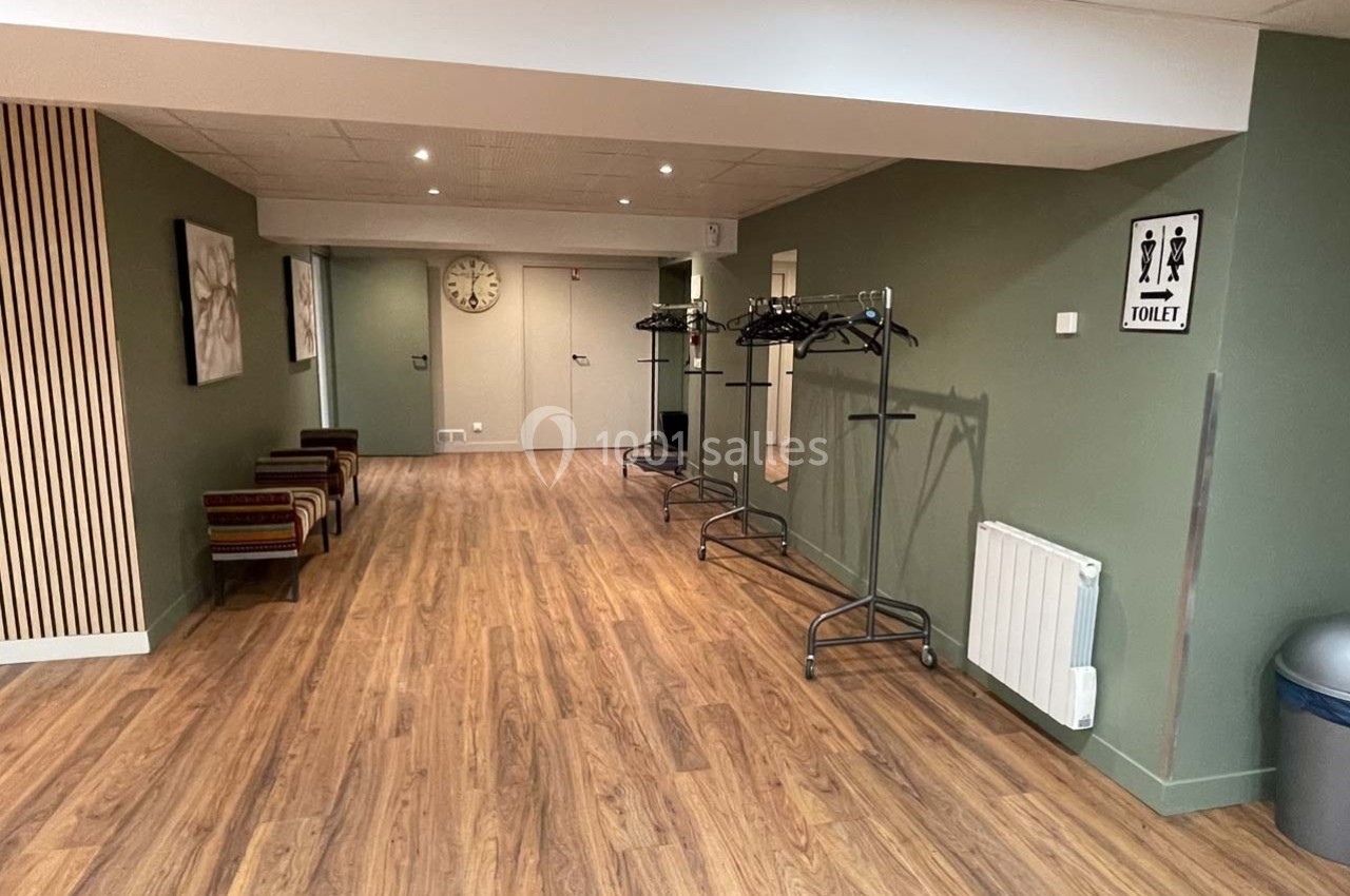 Couloir avec parquet, murs verts, porte au fond, porte-manteaux vides, bancs et panneau indiquant des toilettes.