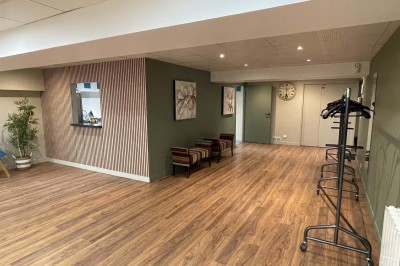 Couloir avec parquet, murs verts, porte au fond, porte-manteaux vides, bancs et panneau indiquant des toilettes.