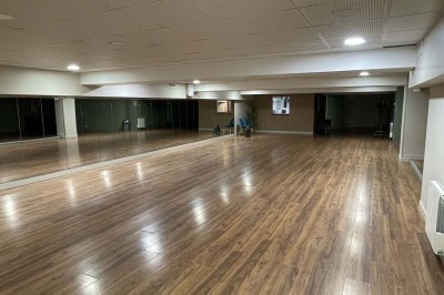 Couloir avec parquet, murs verts, porte au fond, porte-manteaux vides, bancs et panneau indiquant des toilettes.