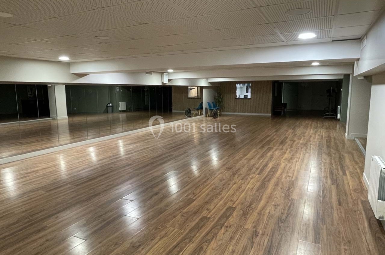 Salle spacieuse avec parquet en bois, murs blancs, miroirs sur un côté et éclairage encastré au plafond.