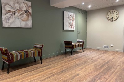 Couloir avec parquet, murs verts, porte au fond, porte-manteaux vides, bancs et panneau indiquant des toilettes.