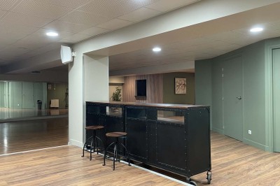 Couloir avec parquet, murs verts, porte au fond, porte-manteaux vides, bancs et panneau indiquant des toilettes.