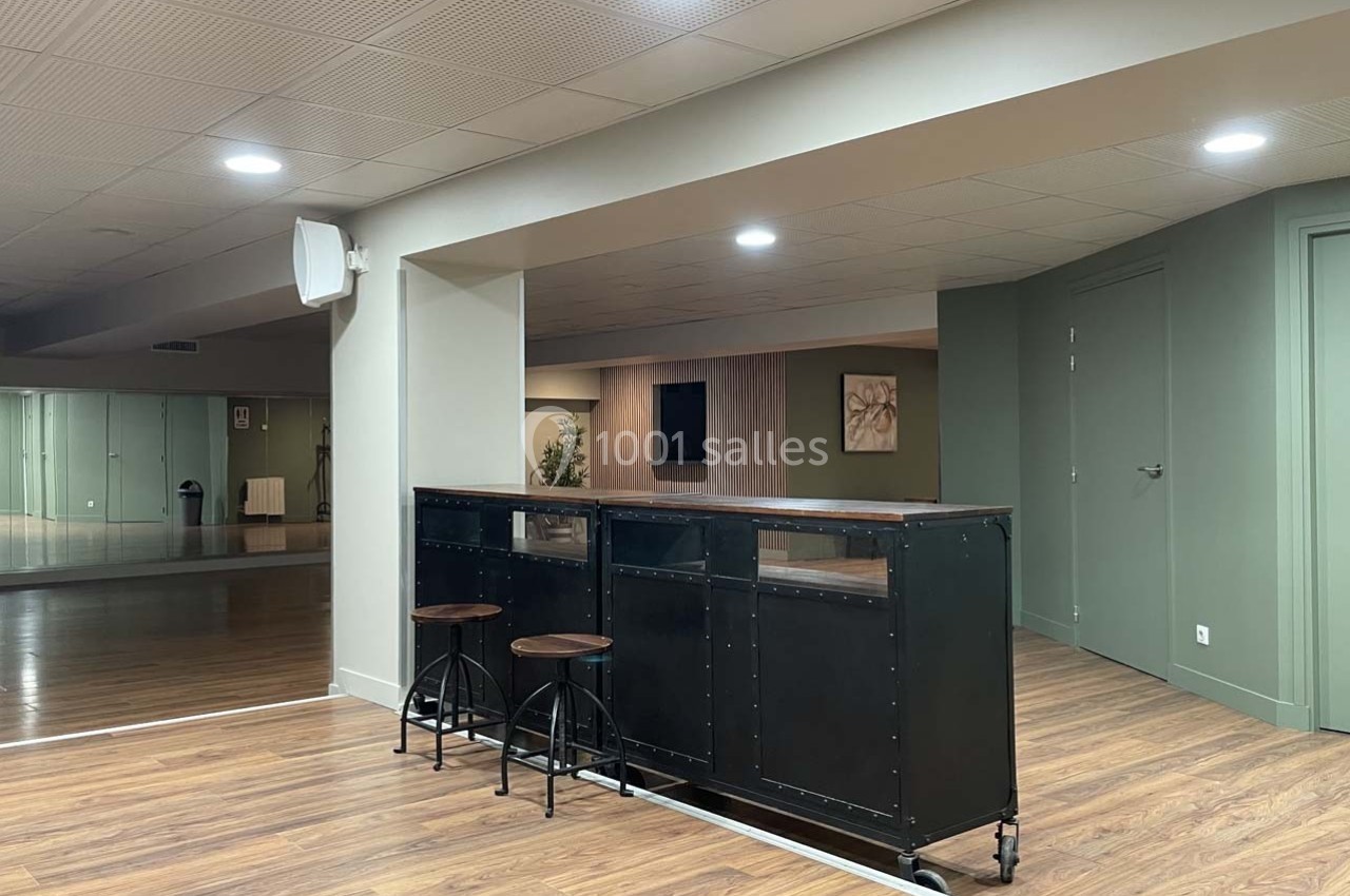 Espace intérieur avec un comptoir noir, deux tabourets hauts, sol en bois et murs verts dans une ambiance lumineuse.