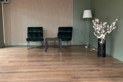 Couloir avec parquet, murs verts, porte au fond, porte-manteaux vides, bancs et panneau indiquant des toilettes.